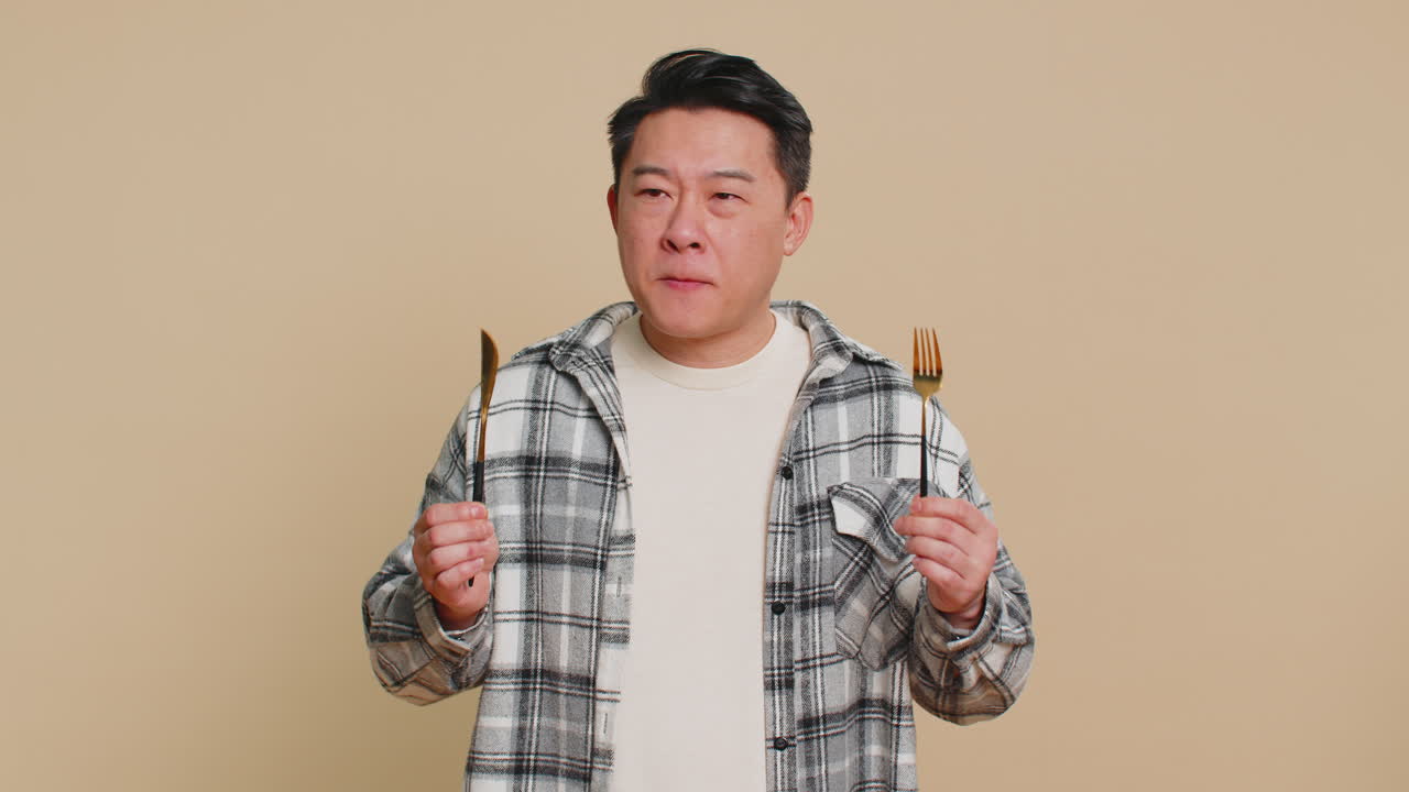 Hungry asian man waiting for serving dinner meal dishes with restlessness holding cutlery fork knife