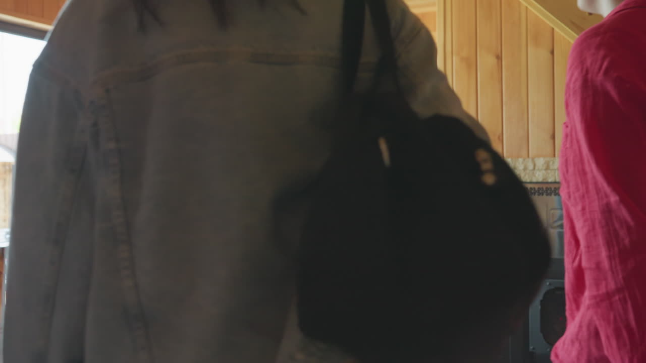 Back view women collecting bags inside cabin kitchen, denim jacket carrying black duffel, red blouse woman with shoulder purse, sunlight streaming through window illuminating rustic ambiance