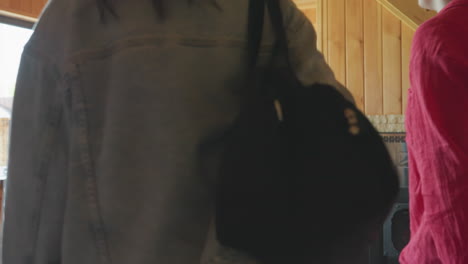 back view women collecting bags inside cabin kitchen, denim jacket carrying black duffel, red blouse woman with shoulder purse, sunlight streaming through window illuminating rustic ambiance