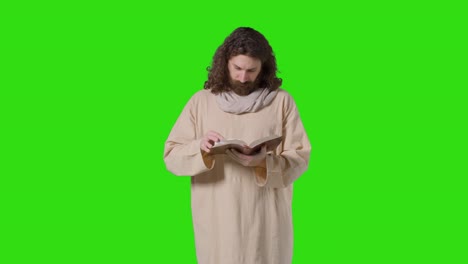 fotografía de estudio de un hombre vestido con túnicas, cabello largo y barba que representa la figura de jesucristo leyendo la biblia en una pantalla verde