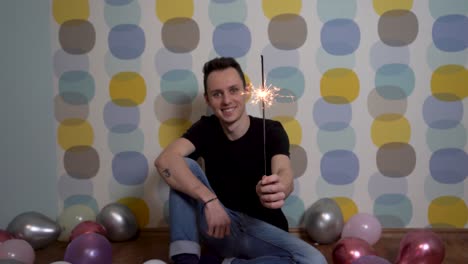 Young-Man-Sitting-with-a-Sparkler-Against-Colorful-Background,-Surrounded-by-Balloons-on-the-Ground