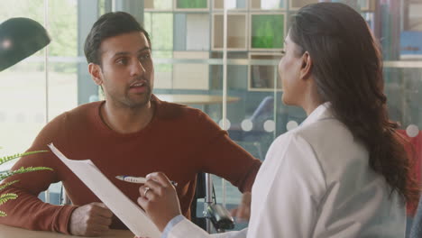 Female-Doctor-In-Office-Discussing-Test-Results-With-Male-Patient-In-Wheelchair-In-Hospital