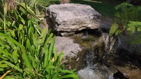 Agua-Dulce-Brotando-De-La-Fuente-Entre-Rocas-De-Granito,-Helechos-Y-Plantas-Decorativas-Junto-A-Usted-En-El-Parque-Paisajístico-En-Una-Soleada-Mañana-De-Verano