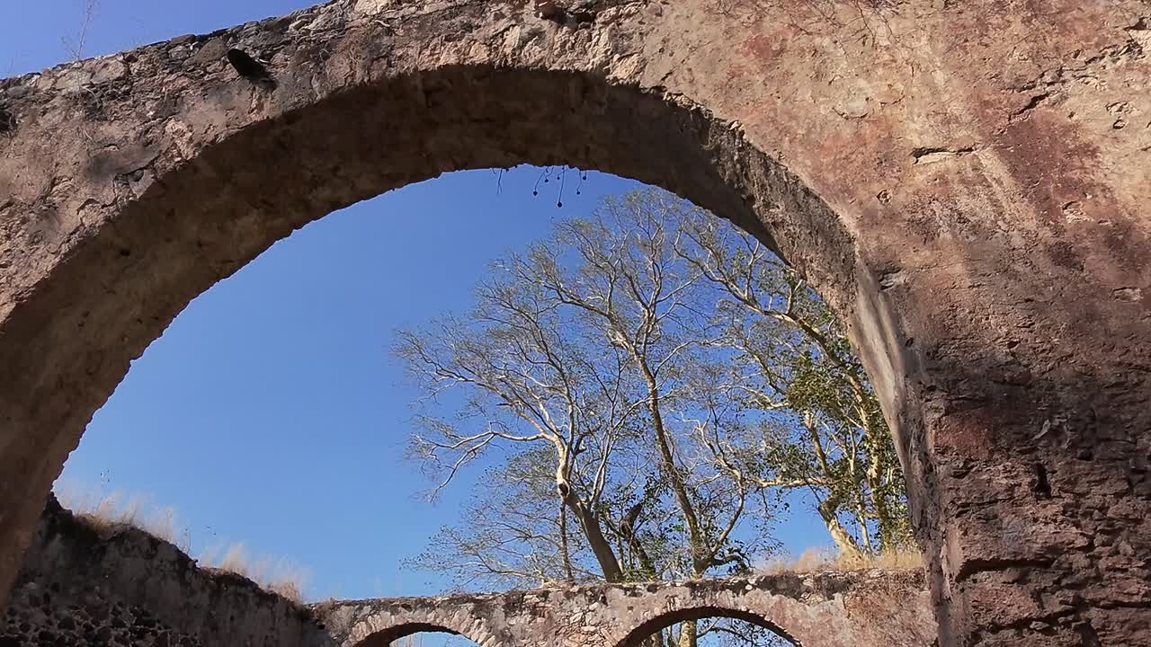 Arched stone ruins at Ex Hacienda San Jacinto Ixtoluca, La Mezquitera, Morelos, with dry trees rising above crumbling stone walls under blue sky. Slow motion tilt down view