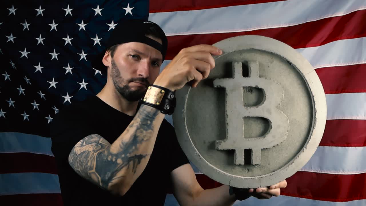 Man with a full sleeve tattoo and black clothing lifting up and holding a large Bitcoin cryptocurrency gray sculpture while standing in front of an American US flag and the man nods with approval.