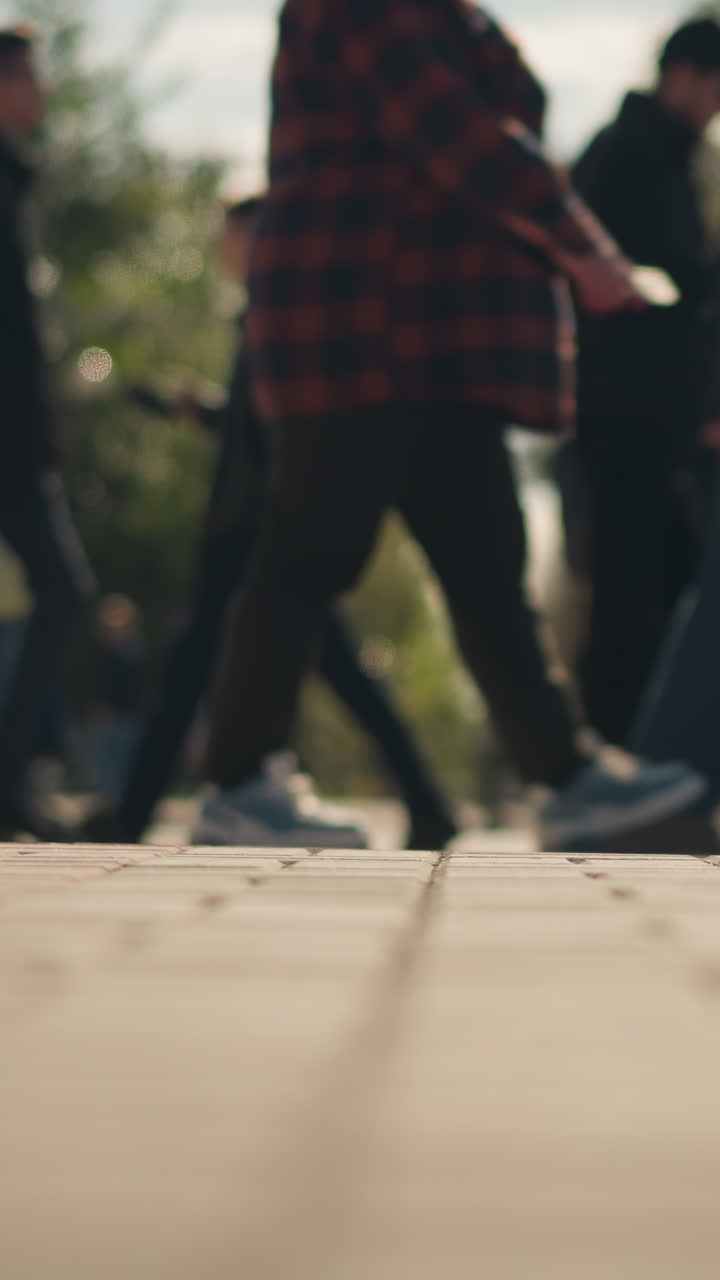 Folla di pedoni che attraversa la strada da una prospettiva dal basso, studenti e pendolari che camminano su un marciapiede pavimentato illuminato dal sole, primi piani di passi e figure sfocate, zaini e abbigliamento casual, alberi autunnali e bokeh caldo, spontaneo