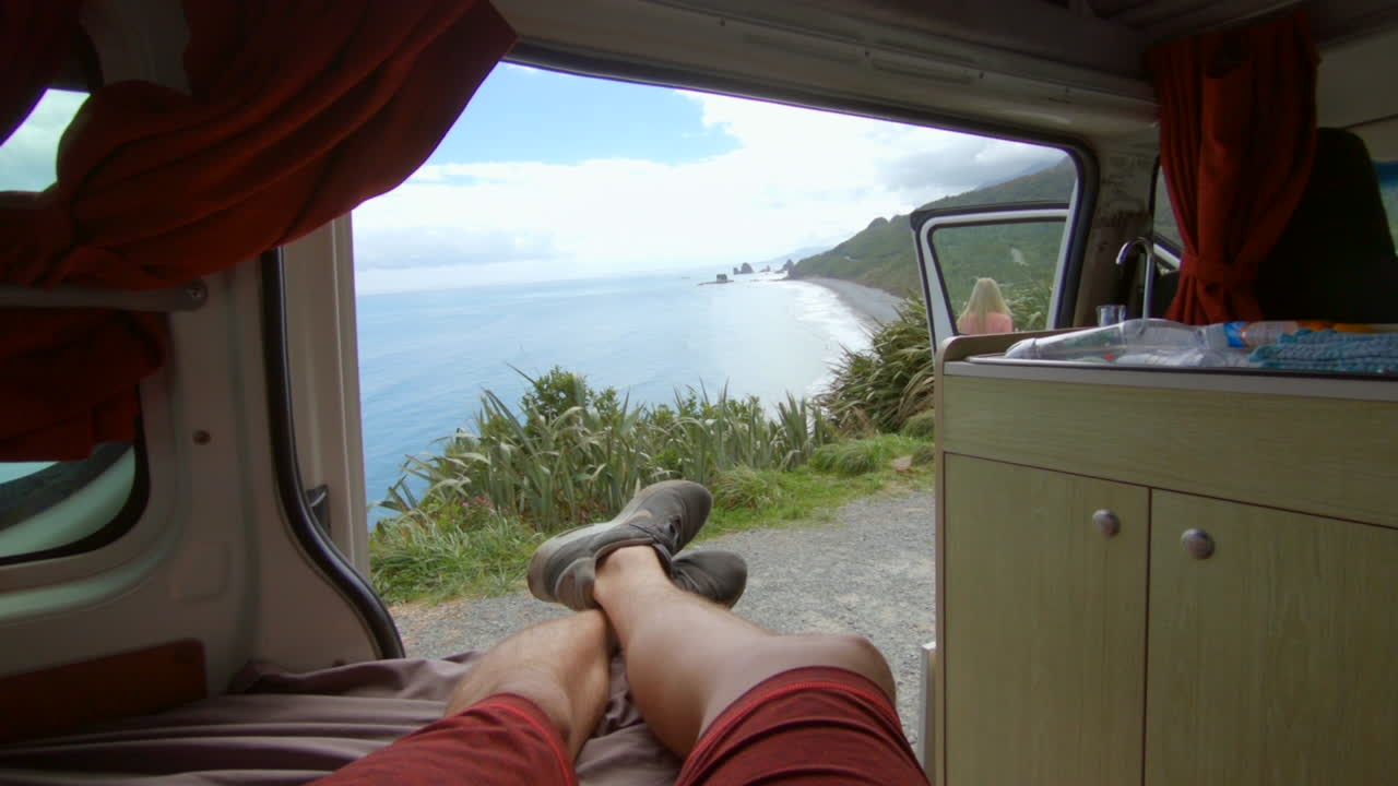 punto de vista de una autocaravana a un impresionante paisaje marino, una chica rubia camina delante del coche, costa oeste, nueva zelanda