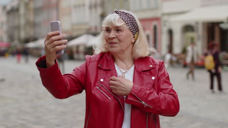 senior woman blogger taking selfie on smartphone video call online with subscribers in city street