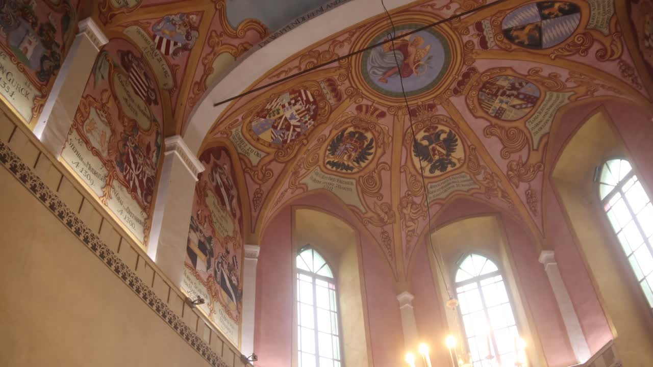 Intricate painted ceiling in historic church with heraldic symbols, detailed artwork, and tall arched windows.