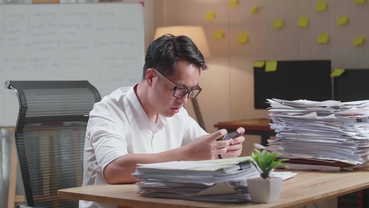 Asian Man Being Upset Losing Game On Smartphone After Working With Documents At The Office