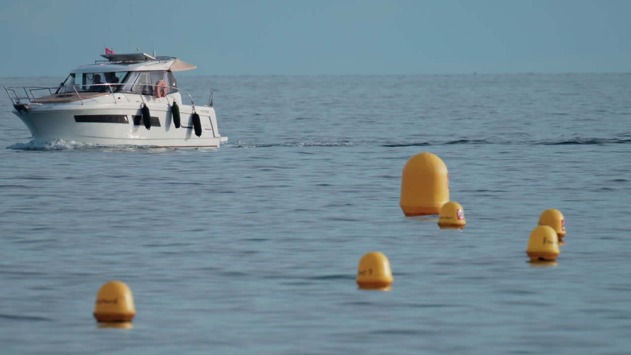 A luxury motor yacht cruises gently across calm blue waters, passing several bright yellow buoys under a clear sky