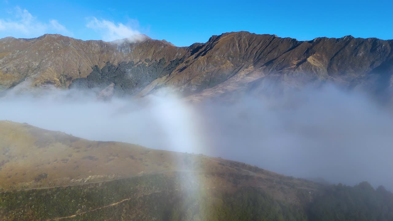 A serene view of a rainbow and fog enveloping the rugged mountains of Moke Lake, Queenstown, under clear blue skies
