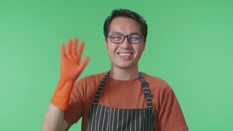 close up of asian male housekeeper with an apron waving hand and smiling while standing in the green screen studio