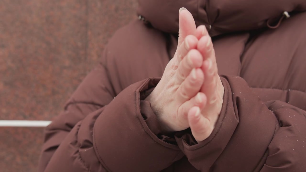 Close-up of adult hands clasping and rubbing together at chest while wearing brown insulated puffer jacket, visible motion of palms and fingers pressing together suggests self-warming in cold weather