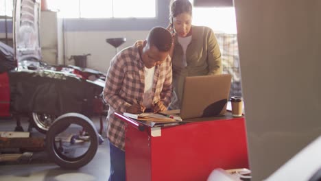 Video-of-two-diverse-female-car-mechanics-using-laptop-and-smartphone,-searching-for-car-parts