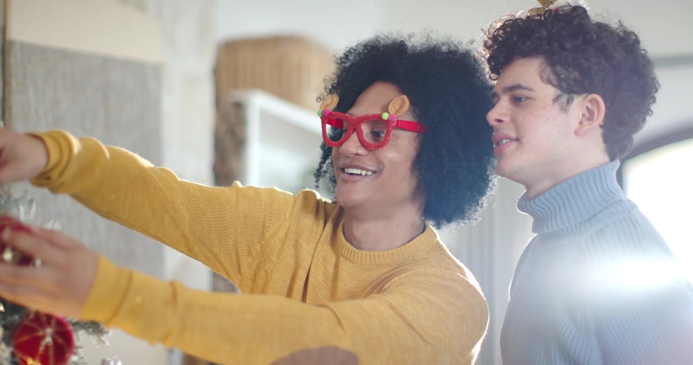 African American men hanging red bauble on tree at home, left in reindeer specs, right pointing
