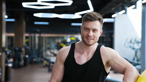 portrait of a sportsman indoors. handsome bearded young athlete in black singlet rests after hard training and looking on camera in gym.