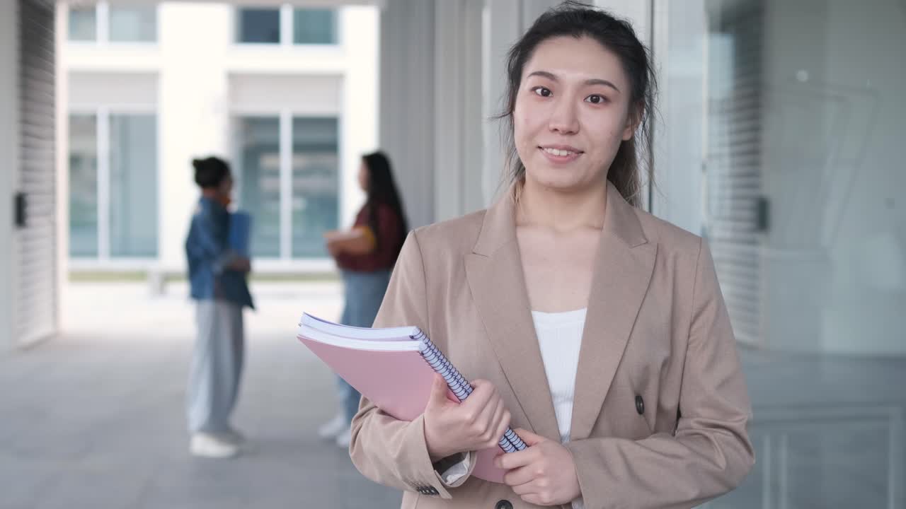 Female university student holding folders and looking at the camera while standing at the hallway. Education concept.
