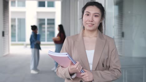 female university student holding folders and looking at the camera while standing at the hallway. education concept.