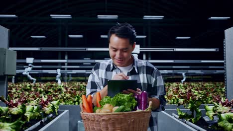 un granjero asiático con una canasta de verduras escribiendo en una tableta en una granja inteligente.