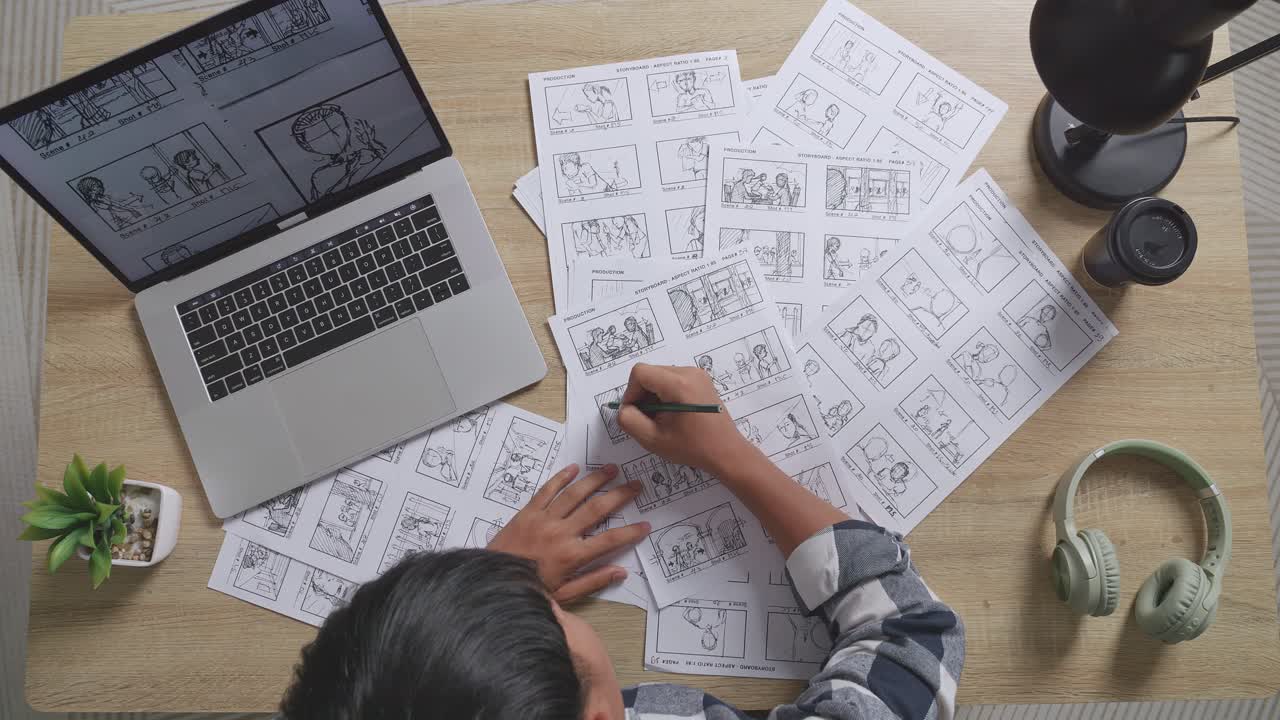 뷰  아웃: 아시안 남성 아티스트가 노트북에서 개념을 스케치하고 스튜디오에서 헤드폰으로 테이블에 스토리보드 그림을 그리는 장면