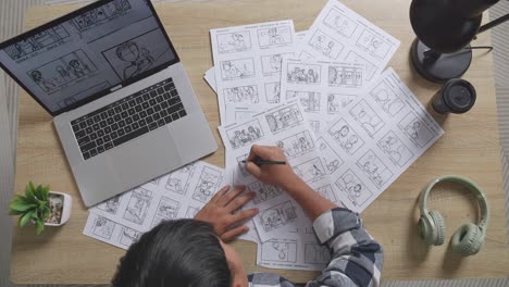 top view zoom out shot of asian male artist looking at sketching concept on a laptop and drawing storyboard on the table with headphones in the studio