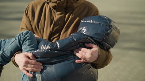 a top to down view of a father carrying his sleeping child while walking, looking stressed, the father wears a brown jacket and beanie, while the child is in a black jacket