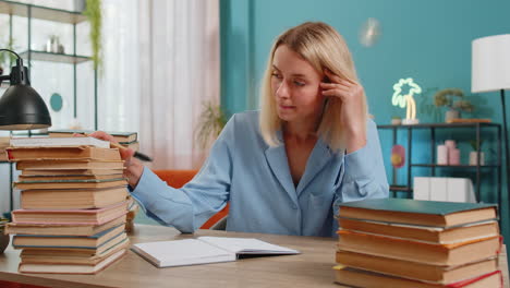 Tired-young-student-writer-woman-sitting-at-desk-with-pile-of-books-writing-a-novel-book-with-pen