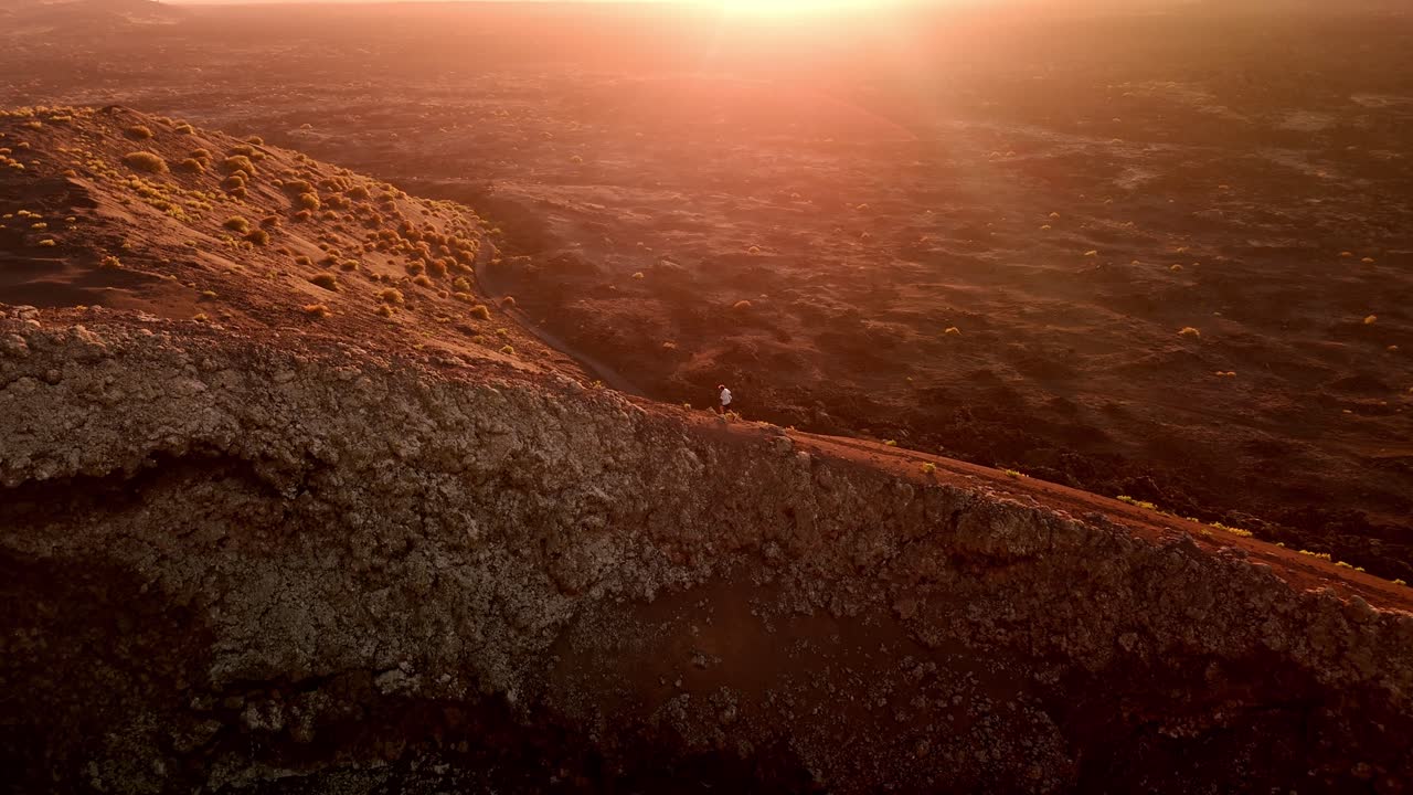 A stunning aerial drone shot capturing a solitary man walking along the rim of Volcano del Cuervo at sunset in Lanzarote, Canary Islands, Spain.