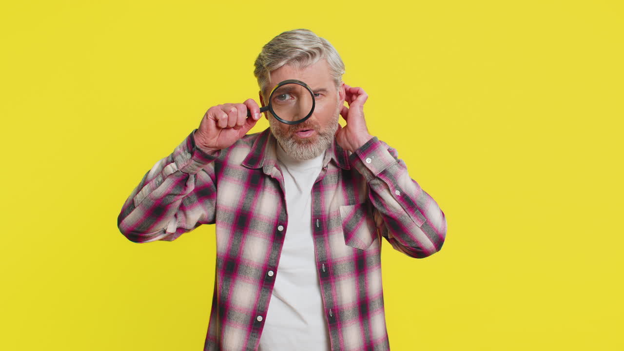 Mature man holds magnifying glass near face looking at camera with big zoomed eye analyzing