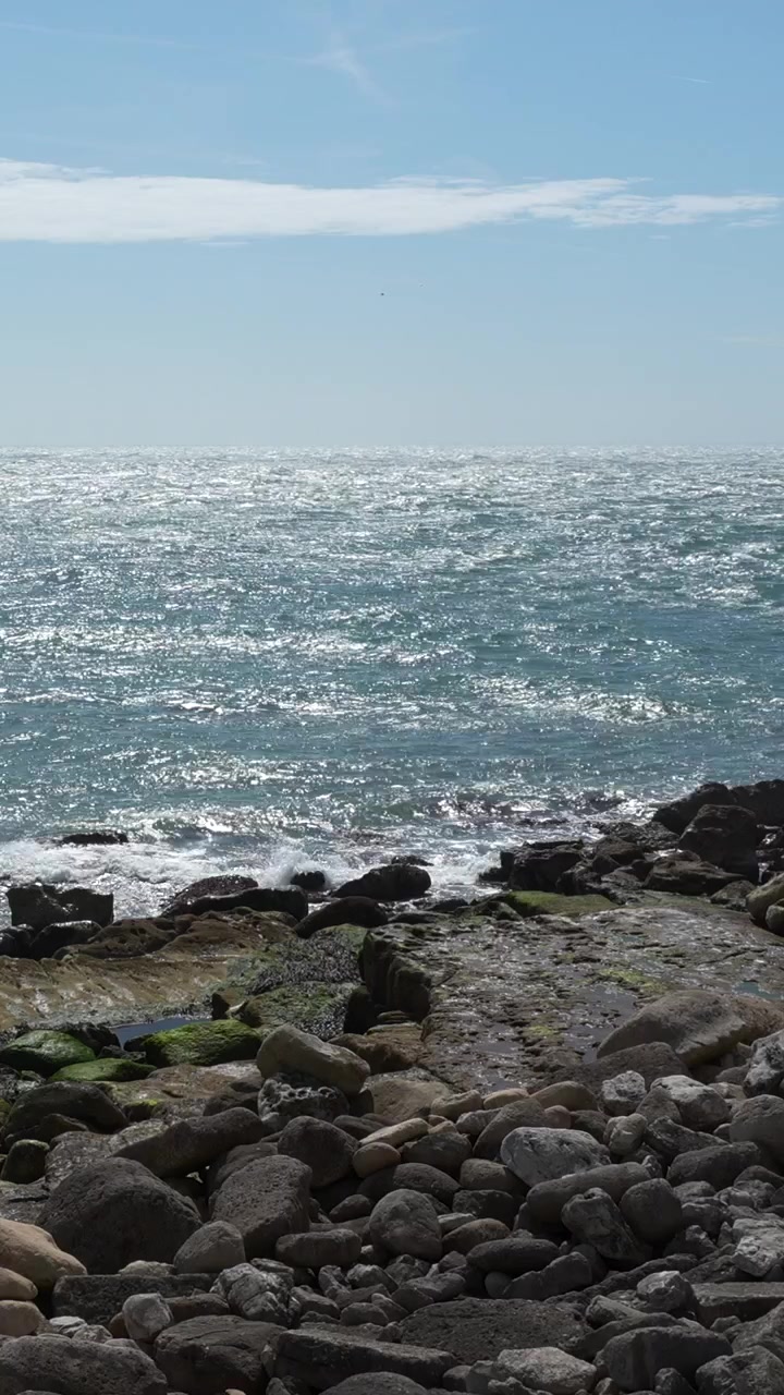 Bidikan vertikal pemandangan sinar matahari yang memantul di air laut biru saat ombak menghantam garis pantai berbatu di bawah langit cerah. Berguna untuk konten perjalanan, alam, dan kesehatan.