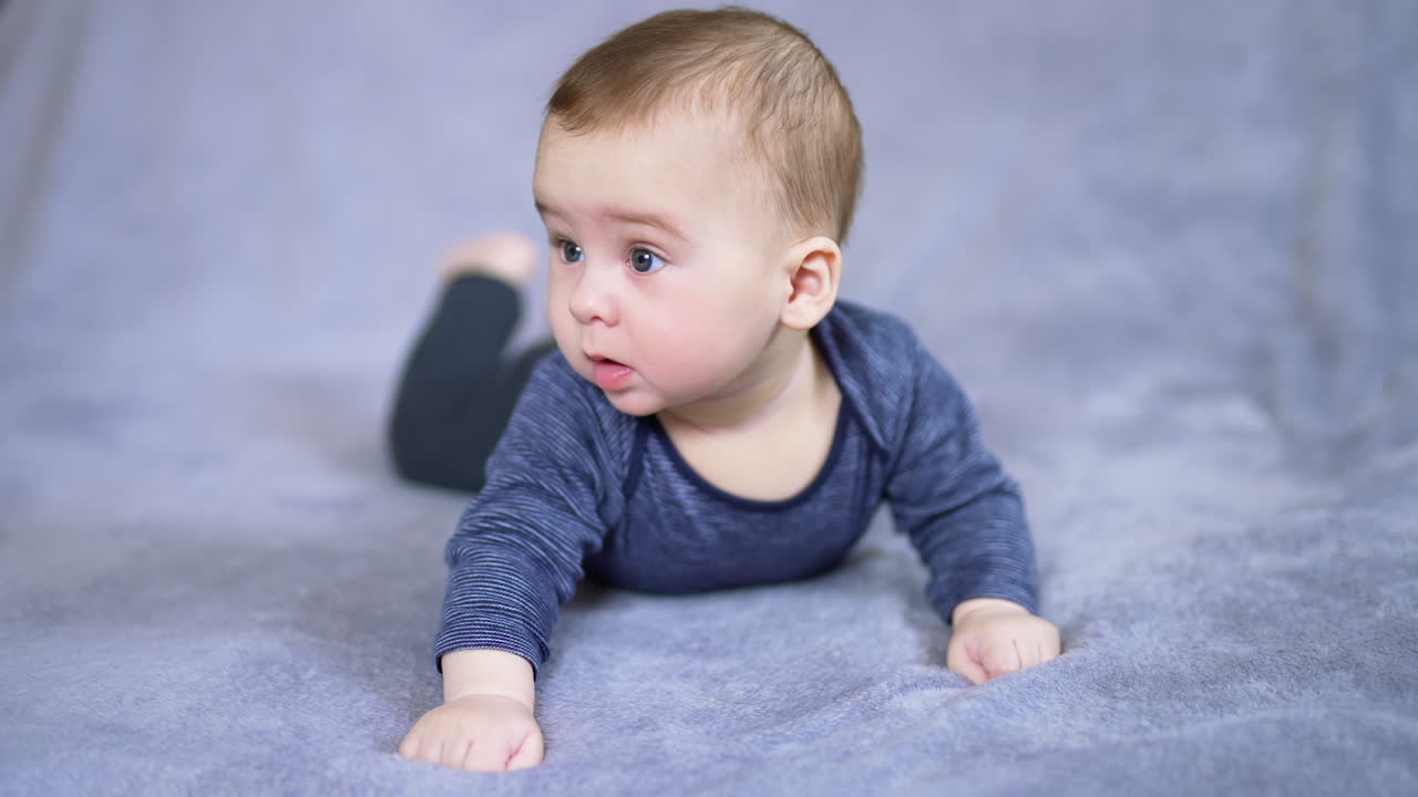 Healthy Caucasian boy lies on tummy and raises himself on arms. Curious toddler looking sideways with interest. Grey backdrop.