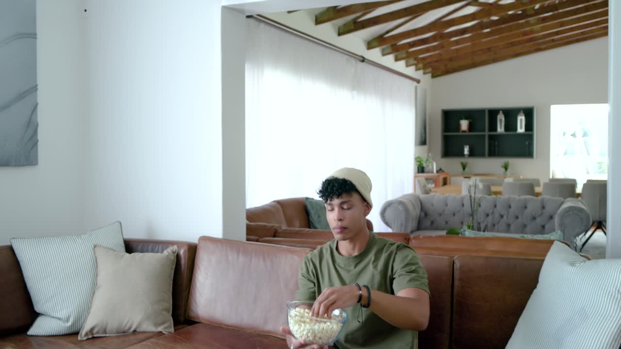 Homem afro-americano adulto segurando uma tigela de vidro com pipoca em um sofá secional marrom, reagindo à TV.