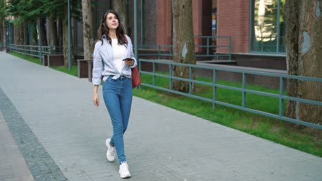 full length view of caucasian student woman wearing headphones and listening to the music on smartphone while walking down the street