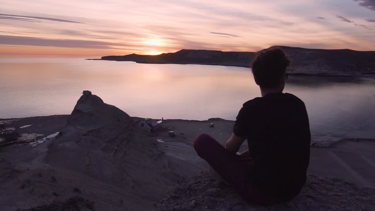 joven modelo sentado en la cima de una montaña viendo la puesta de sol - plano general