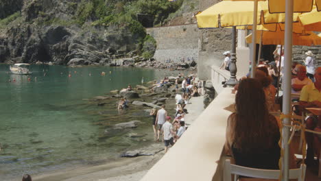 people on the rocky beach and seaside restaurant of vernazza harbor in cinque terre, la spezia province, liguria, italy. sideways shot