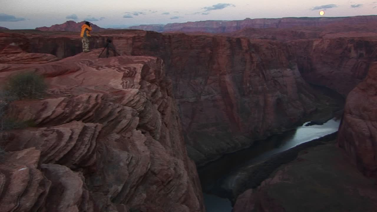 Wide Shot Of A Photographer Standing On Cliff And Photographing The Canyons Of The Colorado River As It Weaves Through Tight Sandstone Canyons Under A Full Moon Rising