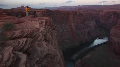 wide shot of a photographer standing on cliff and photographing the canyons of the colorado river as it weaves through tight sandstone canyons under a full moon rising