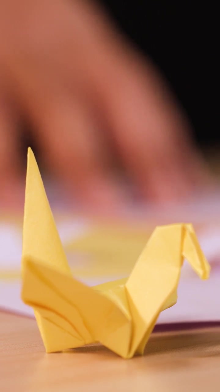 A yellow paper crane sits on a table while hands work in the blurred background