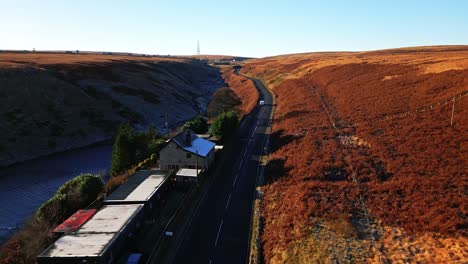 Moving-Aerial-Moorland-View-moving-past-a-farmhouse-on-a-quiet-road-on-Saddleworth-moor,-showing-a-wild-landscape-with-traffic,-reservoir-and-snow-lined-moors