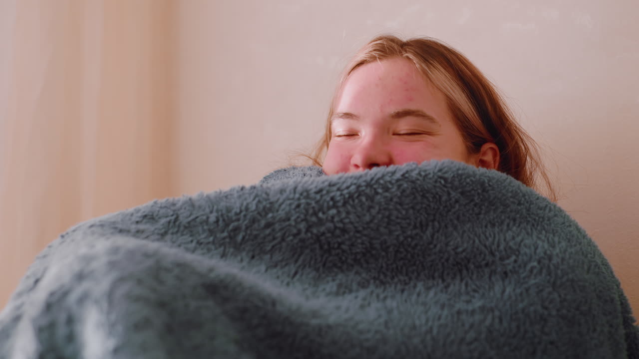 Happy student seated on sofa wrapping herself under blanket enjoying holiday moment with gentle smile as sunlight enters through window creating mood of warmth