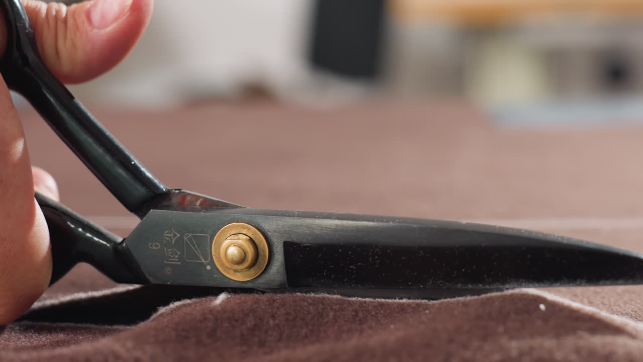 Close up view of hand gripping black scissors cutting brown textile along chalk markings on work surface, precision tailoring process captured in workshop setting, focus on blade slicing fabric
