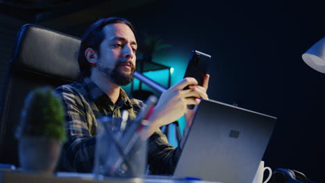 man relaxing in apartment sitting at office desk, texting friends