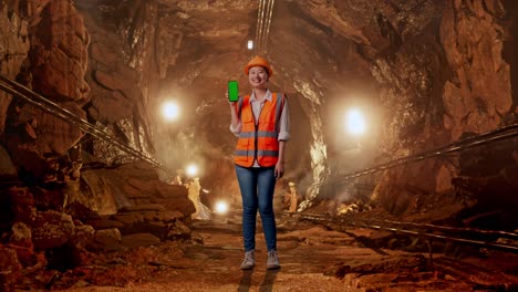 full body of asian female engineer with safety helmet smiling and showing green screen smartphone to the camera in underground mine tunnel