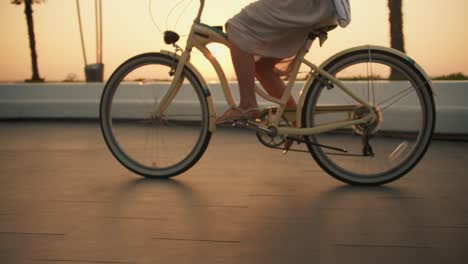 A-girl-in-a-white-dress-pedals-a-bicycle-and-rides-on-the-beach-during-sunrise-near-the-sea.-Morning-bike-ride-to-the-beach-near-the-sea-where-palm-trees-grow