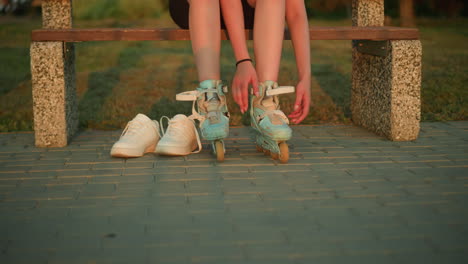 lady removing strap of left leg roller skate while seated on bench in park with two white sneakers by her side, with blurred background featuring greenery