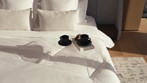 bed side table which includes 2 black coffee mugs and a book on top of a large bed with white bedding