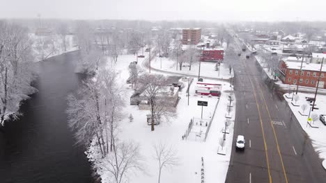 River-near-small-town-street-with-traffic-during-snowfall,-aerial-view