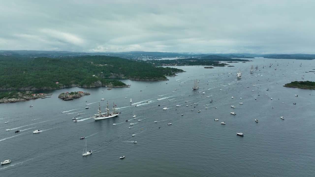 Numerous sailboats and small vessels traverse the waters of a picturesque archipelago under a cloudy sky. The lush green hillsides frame the serene seascape, showcasing nature's beauty