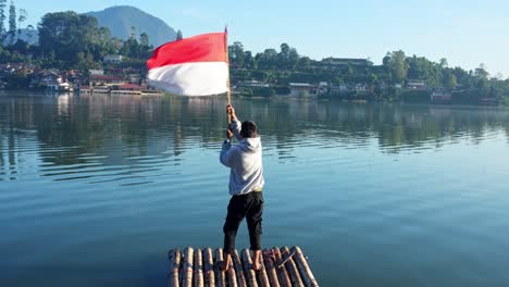 young man proudly holding and waving indonesia flag while standing on bamboo raft with serene lake and reflection